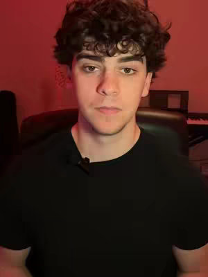 A medium shot of a young man with curly dark hair looking directly at the camera. He is wearing a black t-shirt, and the background is a dimly lit, red-toned space. A microphone is clipped to his shirt. Behind him, a portion of a piano and some studio equipment are visible, slightly out of focus.