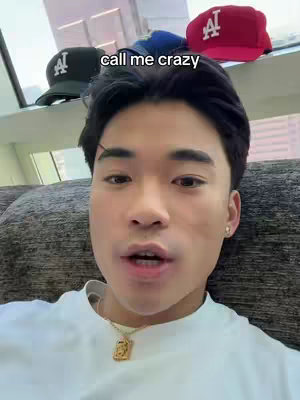 A young man with dark hair is centered in the frame, looking directly at the camera with a slightly open mouth, maybe speaking. He\'s wearing a white T-shirt and a gold chain with a pendant. Behind him, on a shelf, are several baseball caps, including a black one and a red one, both with the letters \"LA\" on them. The background is blurred, hinting at an interior setting, possibly a living room with a grey couch visible on the bottom.