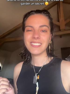 A medium shot of a young woman with dark hair, wet or styled back, smiling at the camera with a backdrop of wooden beams. She wears a black top and various necklaces and earrings.