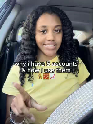 A woman with dark, curly hair is seated in a car, facing the camera. She's wearing a yellow shirt and smiling, with a steering wheel partially in view. Text overlayed on the image reads 'why i have 5 accounts & how i use them,' with a money bag and an upward-trending graph emoji below. The background shows the car's interior.
