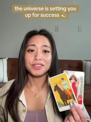 A young woman with long dark hair is holding two tarot cards in front of her. The background includes a light-colored wall, a few pillows, and a hint of a wooden headboard. She's looking at the camera and talking. Overlay text at the top says "the universe is setting you up for success".