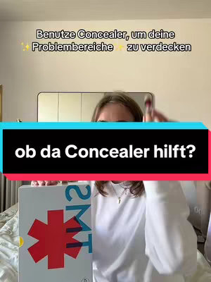 A young woman holds a magazine or notebook with an image of a large red star on it, with text that reads '2MT' and what appears to be a yellow sticker on the lower left corner. She is surrounded by a bedroom setting, with a mirror and bed visible in the background, a phone in her hand, and in front of a bedsheet. The video overlay features german text over a rectangle with a black background. She is dressed in a simple white crew neck top.