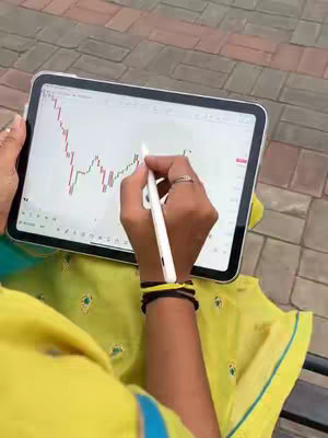 The image shows a person's hands resting on a tablet, angled in a way to show what's on the screen. The background is blurred, but it appears to be an outdoor scene. The screen displays a graph, apparently a stock chart. A white stylus is held over the screen. The person is wearing jewelry and a yellow patterned top with green accents.