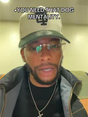 A close-up shot of a Black man with a beard and glasses. He wears a black jacket over a black shirt and a silver cross necklace; a cap with green accents is on his head featuring text in the top left corner. The background is a simple wall.