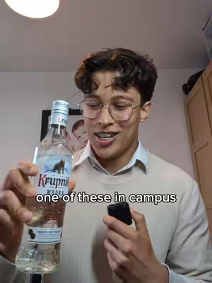 A young man with glasses and braces holds a bottle of vodka and a small black object in front of a wall with some wall decor. The text overlay 'one of these in campus' is centered at the bottom of the image.