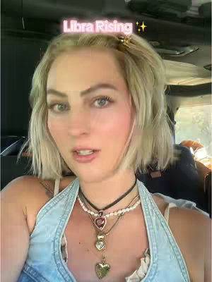 A selfie-style video shows a fair-skinned woman looking towards the camera. She has blonde hair and is wearing a casual outfit and layered necklaces. The background suggests a car interior. Floating at the center-top of the image is overlaid text.