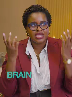 A woman with short, dark curly hair and glasses is speaking, gesturing with her hands. Behind her is a blurry background with a yellow and brown tone. In the foreground is text that says "BRAIN". She is wearing a red blazer, a white button-down shirt, and gold jewelry.