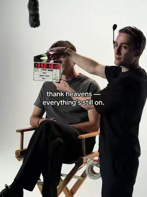 A behind-the-scenes shot of a film or video shoot. Two men are in view: one holding a clapperboard in front of another person seated in a director's chair. The background is a clean white. The image is well-lit and has a professional feel.