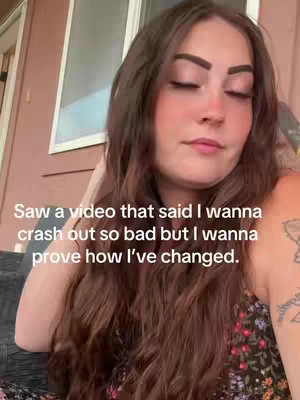 The image shows a woman with long brown hair looking down with her eyes closed. She is wearing a dark, floral top. Text overlayed on the bottom reads, 'Saw a video that said I wanna crash out so bad but I wanna prove how I've changed.' The background is a blurred outdoor setting.