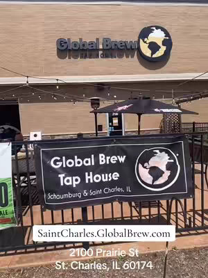The image shows the exterior of a bar or restaurant called Global Brew Tap House. The building has a light brown facade with the name Global Brew in large, dark letters and a circular logo. Below, a banner displays the name and location. In the foreground, a black metal fence and some outdoor seating are visible. The sky is clear and blue, suggesting a sunny day.