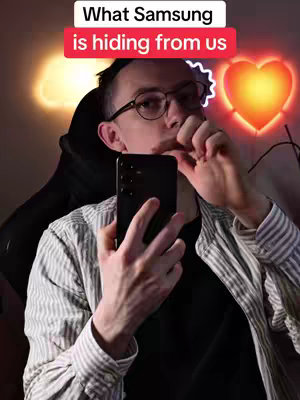 A man with glasses holds a smartphone in front of his face, with one finger near his lips. The background includes an illuminated heart-shaped light and a neon light.