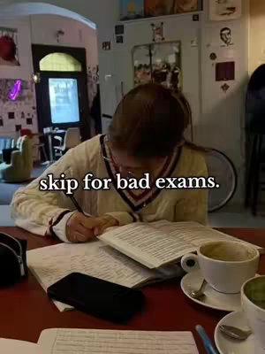 A young woman with glasses is sitting at a table in a cafe and working. She has a book open in front of her and is writing with a pen. There's a cup of coffee to the right of her book, and a smartphone is on the table in front of her. The image is framed from a slightly low angle. The background is a bit blurred, showing a window and some details of the cafe. The lighting is warm and inviting.