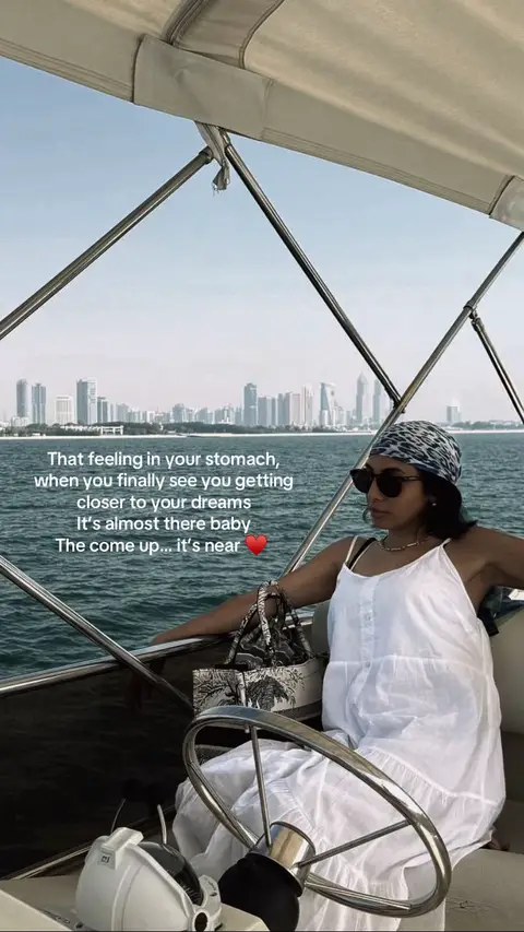 A woman wearing sunglasses and a scarf sits on a boat, with a city skyline in the background. The text overlay reads: "That feeling in your stomach, when you finally see you getting closer to your dreams. It's almost there baby. The come up... it's near" with a red heart emoji.
