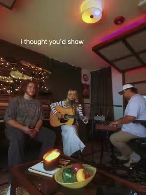 The cover image features three individuals in a dimly lit room with musical instruments and accessories. In the center, a woman with blonde hair is playing an acoustic guitar and singing into a microphone. To her left, a man with long hair is seated, looking at the camera. On the right, a man wearing a cap is sitting at a keyboard, playing as well. In the foreground, a table holds a lit candle and a bowl of fruit. String lights and other decorative items are visible around the room. The overall tone is warm and intimate.