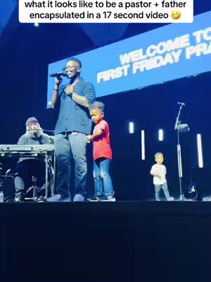 The image shows a man speaking into a microphone on stage with two young children, a musician behind a keyboard, and a banner that reads 'WELCOME TO FIRST FRIDAY PRA'. The man is wearing a black shirt and jeans. One child is standing next to him wearing a red shirt and jeans, and another is standing further back on the stage. The stage has lighting elements on the back wall, and the overall setting appears to be a church or event venue.