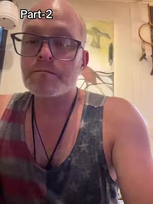 A bald man with glasses and a beard is in a selfie-style video. He's wearing a tank top with a stars and stripes design, and a black necklace is visible. The background is a bit cluttered, suggesting an interior setting.