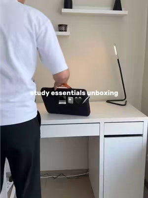 A person standing behind a white desk, unboxing a black laptop bag. The desk has a lamp and some shelves with decorations. The text overlay reads 'study essentials unboxing'.