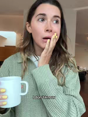 A woman in a green sweater looking to the side with a surprised expression, her hand covering her mouth. She is holding a white mug. The background is a blurred interior.