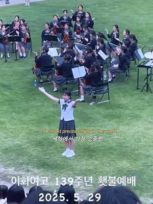 A wide shot of a green grassy field with several groups of people. In the foreground, a young woman in a white shirt, blazer, and skirt stands with arms outstretched. Behind her, a group of young women in similar school uniforms are seated. Behind the young women are other figures seated. The image has text overlays on the grass and at the bottom of the view.