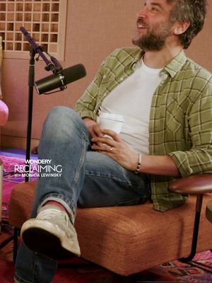 A man sits on an orange upholstered chair with a microphone in front of him, holding a white cup. He wears a green plaid shirt over a white T-shirt, jeans, and grey and white sneakers. The background is a soft, blurred indoor setting and a logo is on the lower left corner.