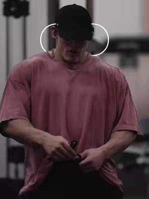 A muscular man in a pink t-shirt and black baseball cap adjusts a strap in a gym. The background is blurred, suggesting an active environment like a gym.