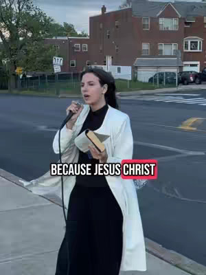 A woman stands outdoors holding a microphone and a book, addressing the camera. A text overlay reads "BECAUSE JESUS CHRIST" in a red rectangle. The background shows buildings and a street.