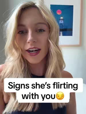A young woman with long blonde hair is centered in the frame, smiling and looking directly at the viewer. Overlayed on the image is a white rectangular box containing the text 'Signs she's flirting with you' in a bold black font. Underneath the text is a smiley face emoji.