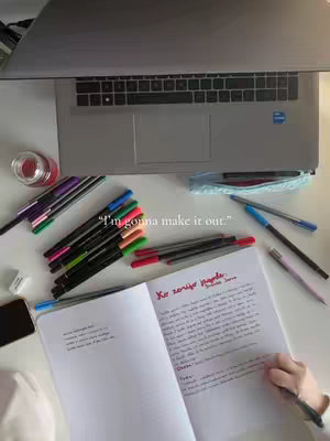 A top-down shot displays a white surface with a laptop at the top. Below the laptop, there are multiple colored pens scattered around an open notebook. In the notebook's right-hand page, the writing is visible. A phone is on the bottom left and some other objects like an ink bottle can also be seen.
