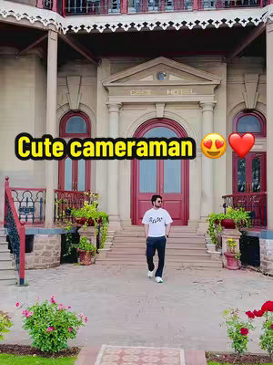 A man is walking towards the camera on a pathway in front of a large building with columns, red doors, and decorative plants. Text overlays highlight the man and the context.