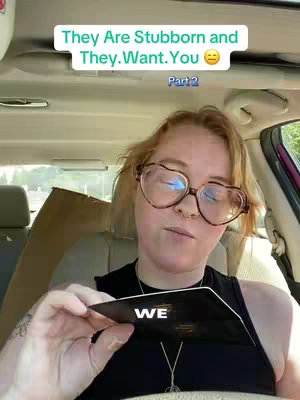 A woman with red hair and glasses is sitting in a car, holding a black card that says "WE". There is overlaid text at the top.