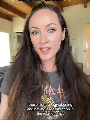 A woman with long brown hair and blue eyes faces the camera in an interior setting. She appears to be mid-sentence with a gentle expression. Text overlays indicating "Awakening to Love" at the top are present, and in the bottom a white text with the message "these signs are awakening and having a recalibration in the relationships"