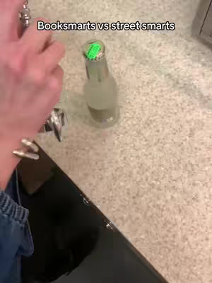 A person's hand pouring liquid from a shiny metal tool into what appears to be a soda bottle, with the label not visible. The bottle sits on a light tan, speckled surface, possibly a countertop. In the background out of focus is a cabinet.