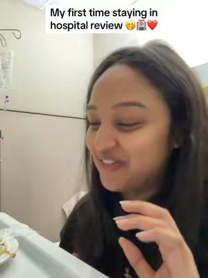 A selfie-style video shows a young woman sitting in a hospital room, presumably recording a review of her experience. The woman, who appears to be in her 20s, is looking towards the side and talking. Behind her, medical equipment such as IV drips is visible, clearly indicating a hospital setting. The room has a neutral color palette with white walls and a reflective surface to the left of the frame. Additionally, a tray with food elements suggests recent use.