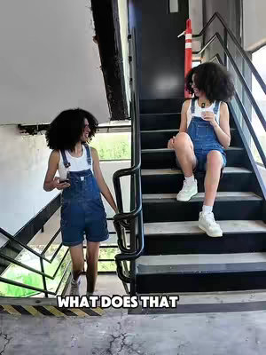 The image shows two young women, resembling each other, in short denim overalls and white tank tops, positioned on a flight of stairs against a backdrop of a building with dark accents and a window. One woman is walking down the stairs holding a phone, while the other is sitting on the stairs, holding a cup. Text reading 'WHAT DOES THAT' is overlaid on the bottom of the image. The overall composition is casual and somewhat artistic.