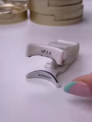 The image shows a close-up of a white eyelash curler on a white surface. The curler is oriented towards the viewer and has "UP" and "DOWN" labels. The background features elements that appear to be makeup containers.