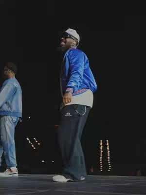 A man in a blue satin jacket and black pants is seen from the side on a stage with one other person visible. The man is smiling with a hat and sunglasses, facing to the right. The background is dark with some lights.