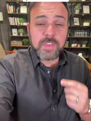 A man with a beard and dark hair looks at the camera while pointing to his right. He wears a black button-down shirt, and there are shelves with items and plants in the background.