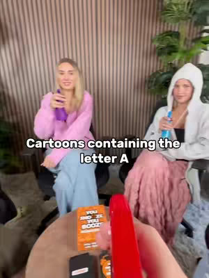 Two young women are seated in a room, holding bottles. A hand holding a red object is in the foreground. Text overlaying the image says 'Cartoons containing the letter A'.