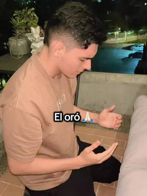 A young man in a beige shirt kneels on a terrace, gazing downward with hands open. Text "El oro" is overlaid. The setting is outdoors with trees and a glimpse of a car in the background.