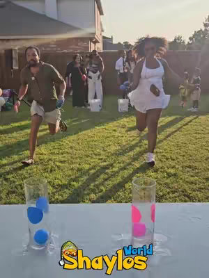 Two people running in a backyard towards the camera, part of a game with glasses filled with water sitting on a table. There are several other people in the background.