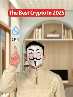 A person wearing a Guy Fawkes mask, posing in front of a blurred background of a room. Above the person is a red banner that says 'The Best Crypto In 2025'. The person is giving a high-five with the index finger.