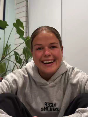 A young woman with fair skin smiles broadly at the camera, her eyes crinkling at the edges. She wears a gray hoodie with the word "KEEP SMILING" printed in reverse across the chest. The background suggests an interior setting, with a glimpse of a plant and part of a wall.