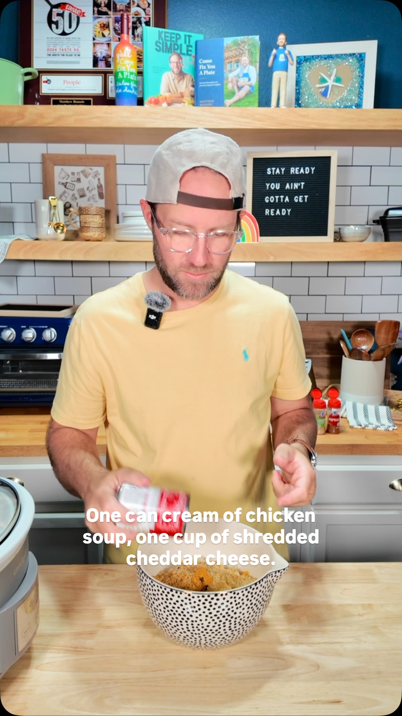 A man wearing glasses, a hat, and a yellow shirt is in a kitchen preparing a meal. In the center is a bowl of cheesy dish with a can of soup next to it and instructions written on the screen.