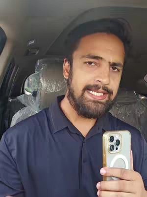 A man with a beard is seated in a car, holding up a phone. He has a slight smile and is looking directly at the camera.