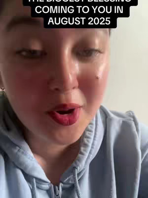 A close-up shot of a person's face, looking toward the camera with red lips and a light blue hoodie. The top part of the image is black with the words 'The Biggest Blessing Coming to You in August 2025'.