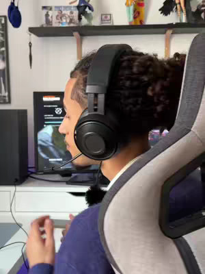 A man wearing headphones is seated in front of a computer screen, with shelves visible in the background. The image has a medium shot composition, showing the man's side profile. The color scheme involves muted tones, providing a clean and focused look.