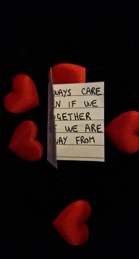 A black background with the words "ALWAYS CARE EVEN IF WE ARE AWAY FROM" written on a folded white piece of paper in black ink. There are four red heart-shaped objects placed around the paper.