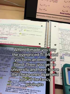 The image is taken from a first-person perspective, with a notebook and a TI-84 calculator in view. The notebook is open to a page filled with handwritten notes. A calculator and more notebook pages are visible on the right side. The background includes what appears to be a table or desk surface, and is a mix of colors.