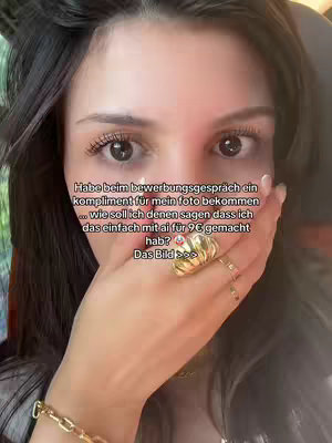A selfie-style shot featuring a young woman with dark hair, staring intently at the camera. She's partially covering her mouth with her hand, wearing a golden ring. The background is blurred, and overlaid text in white describes a compliment received about her photo during a job interview.
