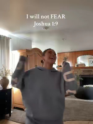 A woman is in a room, raising her arms and smiling. Text overlays state "I will not FEAR Joshua 1:9". The background is a living room setting with wooden furniture.